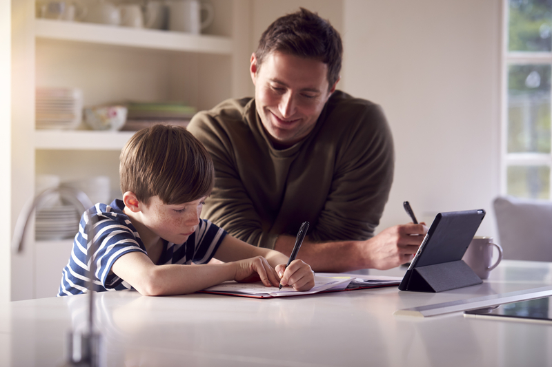 Father Helping Son With Homework Sitting At Kitchen Counter Using Digital Tablet Together